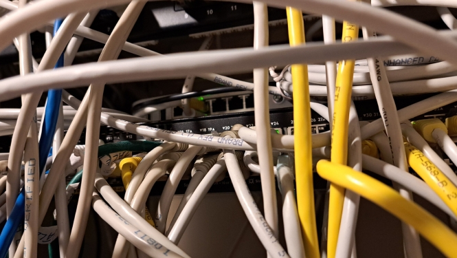 Tuvplānā attēlots sakaru skapis ar lielu daudzumu savstarpēji savijušos tīkla kabeļu. Baltie, dzeltenie un zilie Ethernet kabeļi aizsedz tīkla iekārtas, taču fonā redzams komutators ar vairākiem pieslēgumiem un zaļiem darbības indikatoriem. Attēls ilustrē sarežģītu un grūti pārskatāmu kabeļu organizāciju.