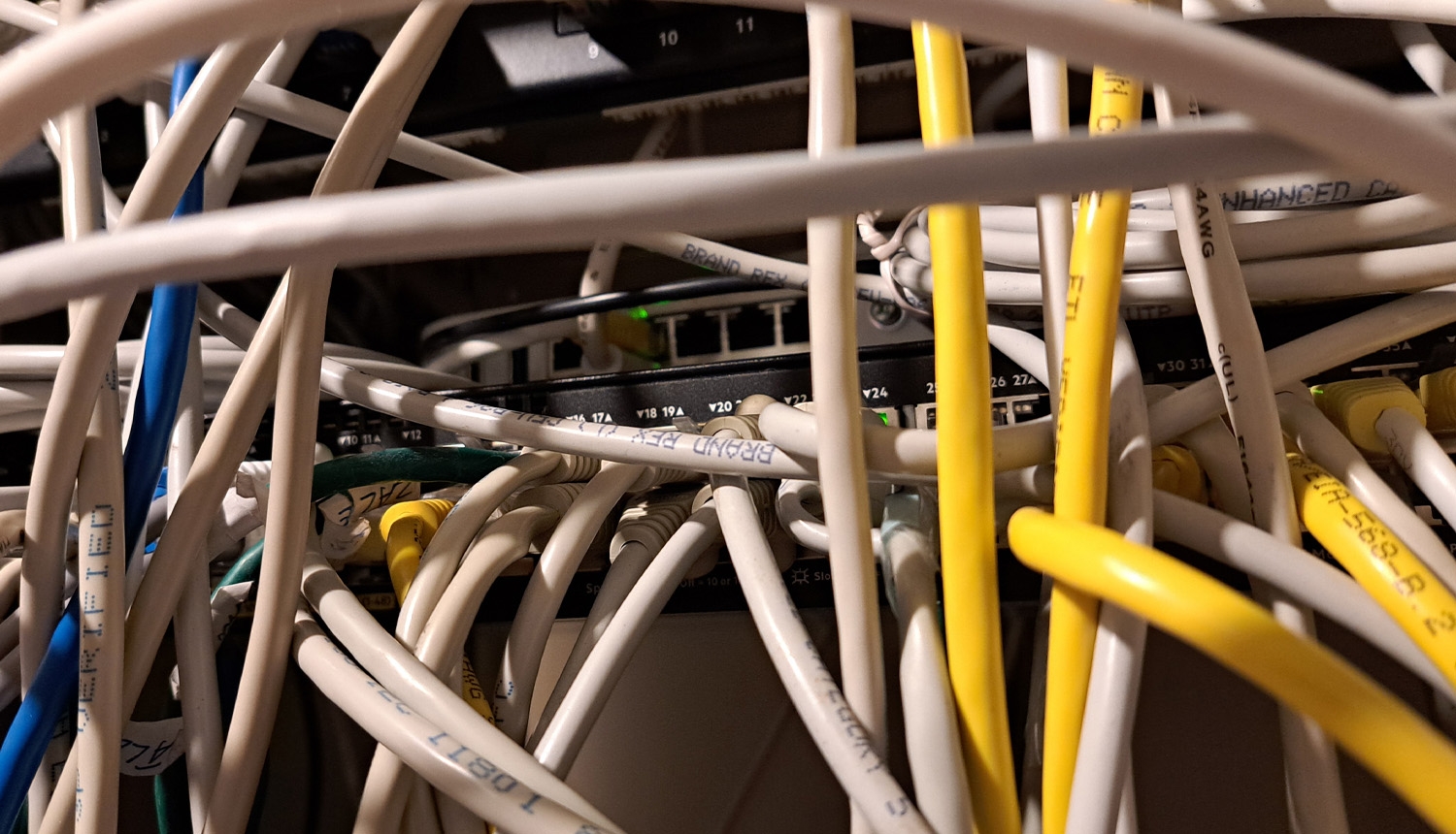 Tuvplānā attēlots sakaru skapis ar lielu daudzumu savstarpēji savijušos tīkla kabeļu. Baltie, dzeltenie un zilie Ethernet kabeļi aizsedz tīkla iekārtas, taču fonā redzams komutators ar vairākiem pieslēgumiem un zaļiem darbības indikatoriem. Attēls ilustrē sarežģītu un grūti pārskatāmu kabeļu organizāciju.