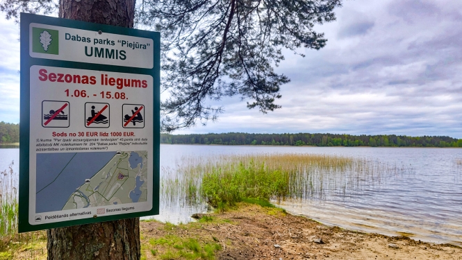 Ezera krasts, apmākusies diena. Informācijas zīme par peldēšanās sezonas liegumu 01.06.-15.08.