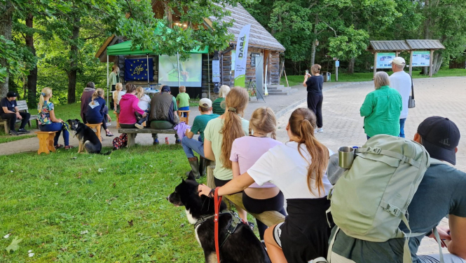 Cilvēli sēž uz soliņiem un klausās stāstījumu.