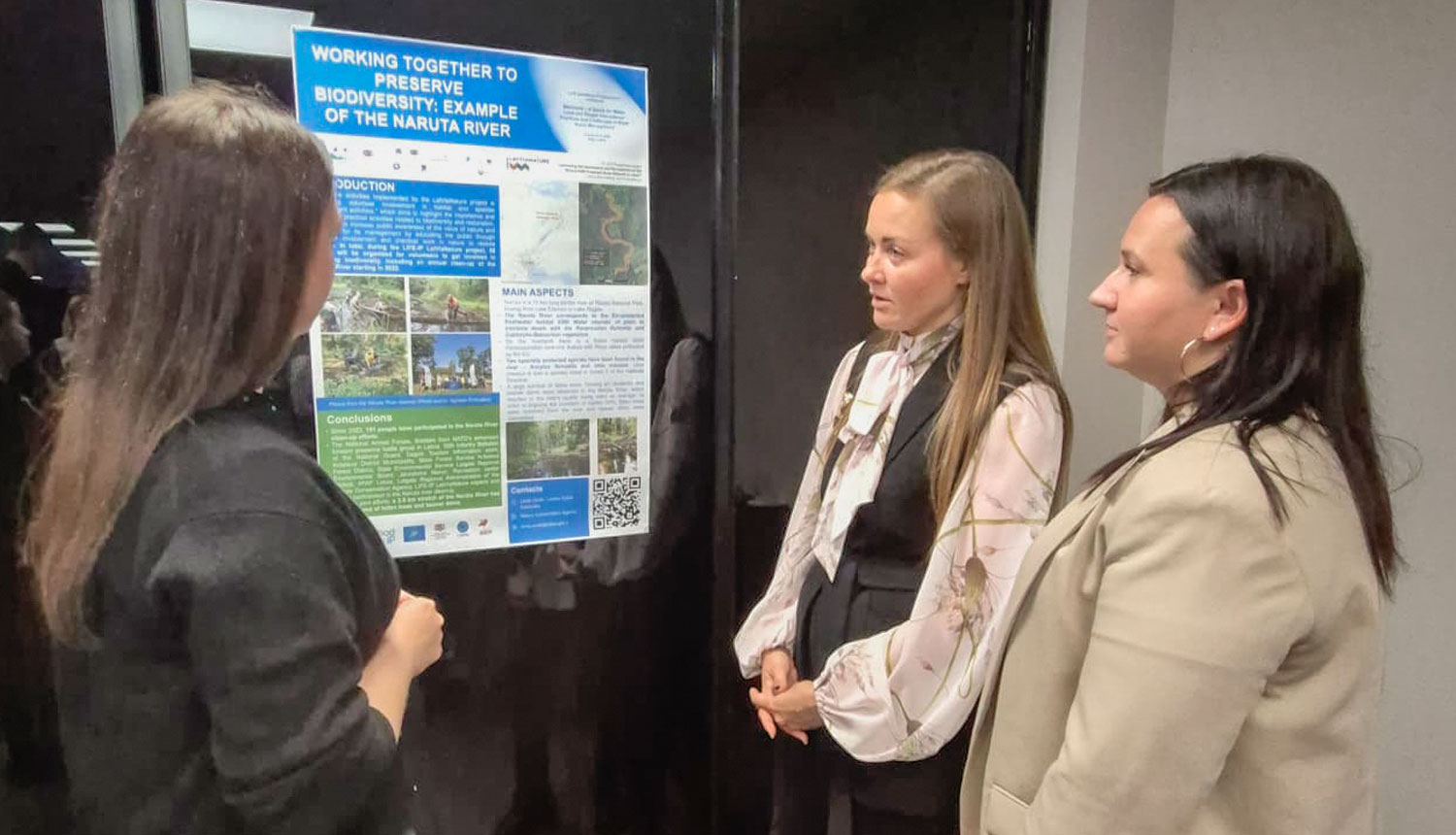 Trīs cilvēki apspriežas pie stenda referāta